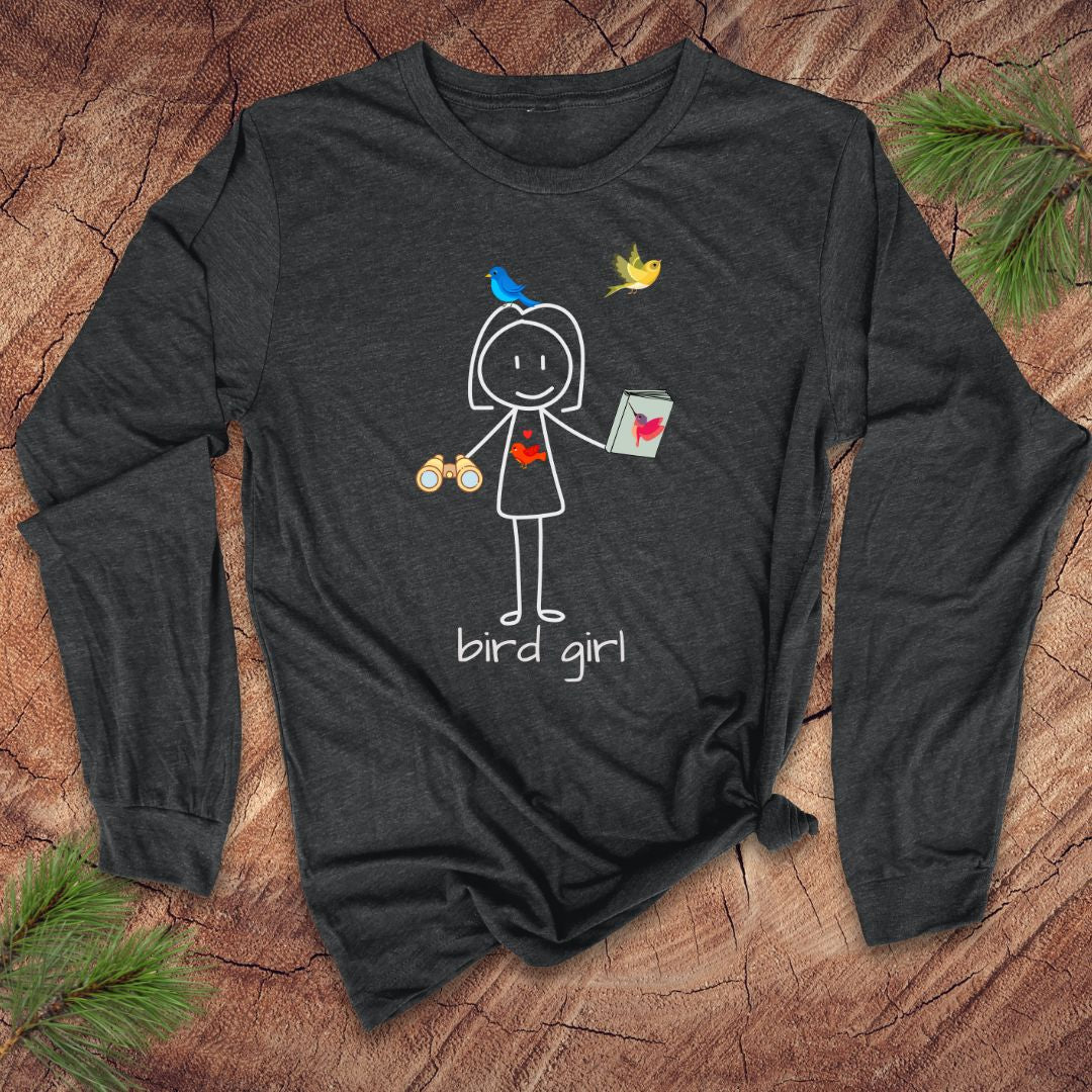 Dark grey long-sleeve shirt with 'bird girl' text and stick figure girl with binoculars and guide book, on a wooden surface with pine branches.