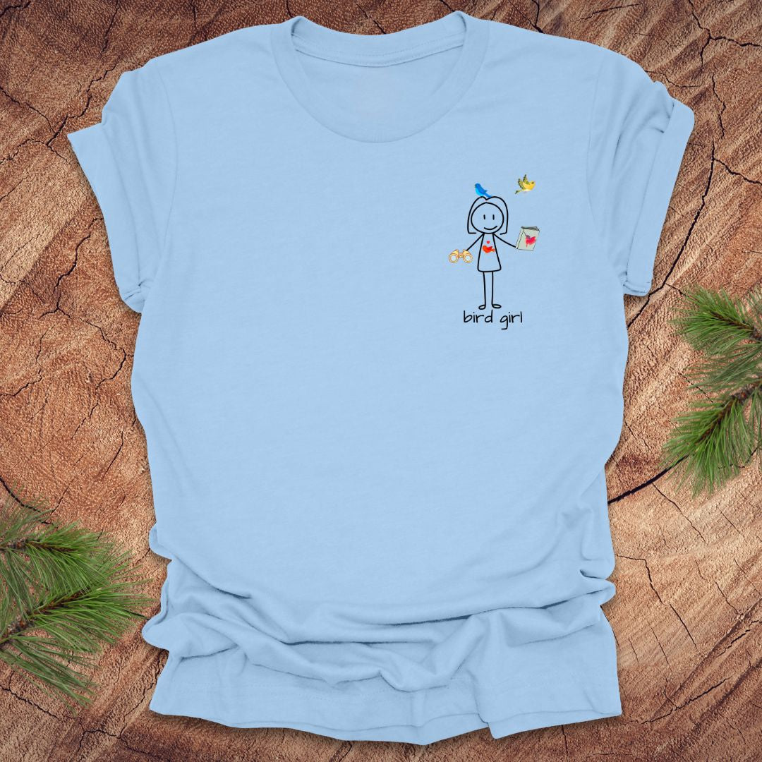 Baby blue t-shirt with a small stick figure and text, 'Bird Girl' on the left pocket side on a wooden surface