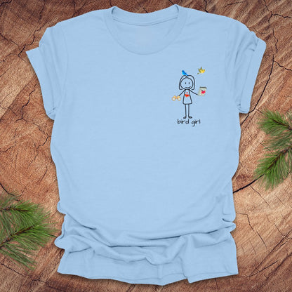 Baby blue t-shirt with a small stick figure and text, 'Bird Girl' on the left pocket side on a wooden surface