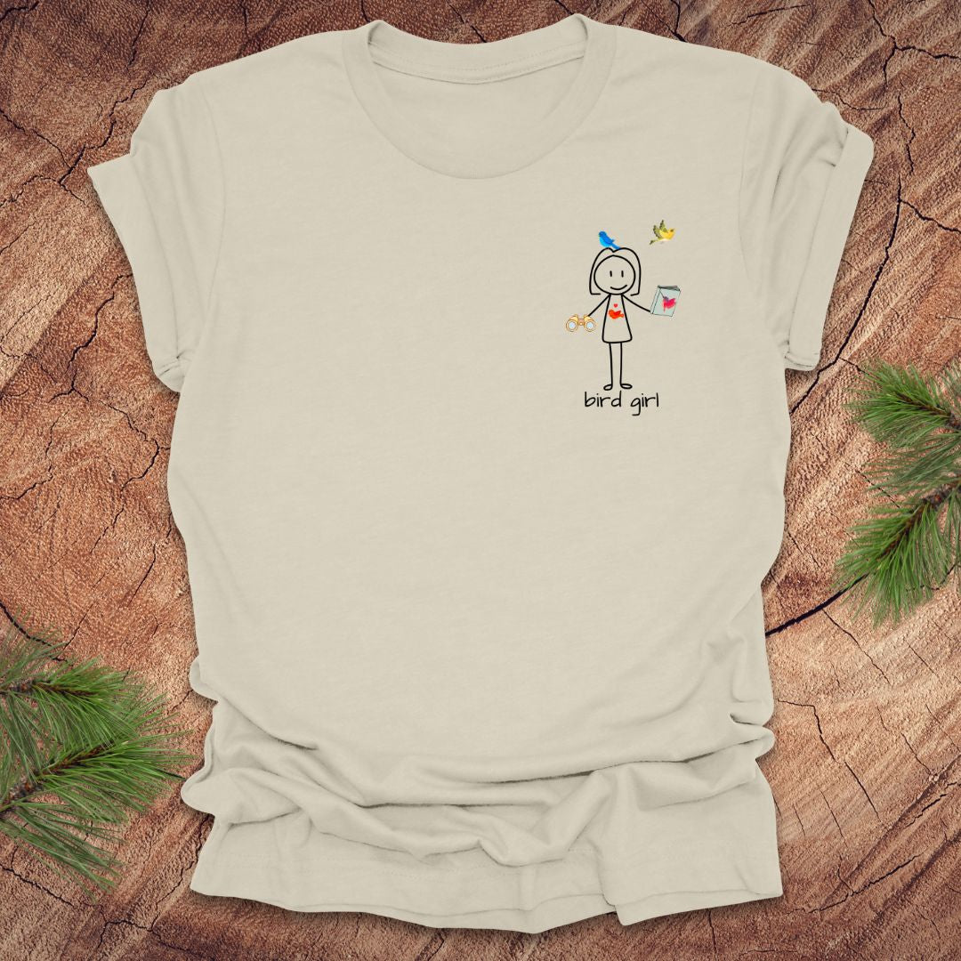 Beige t-shirt with a small stick figure and text, 'Bird Girl' on the left pocket side on a wooden surface