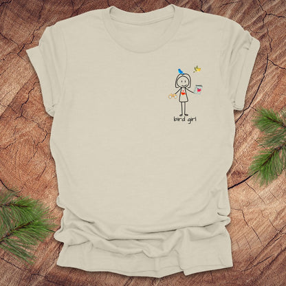 Beige t-shirt with a small stick figure and text, 'Bird Girl' on the left pocket side on a wooden surface