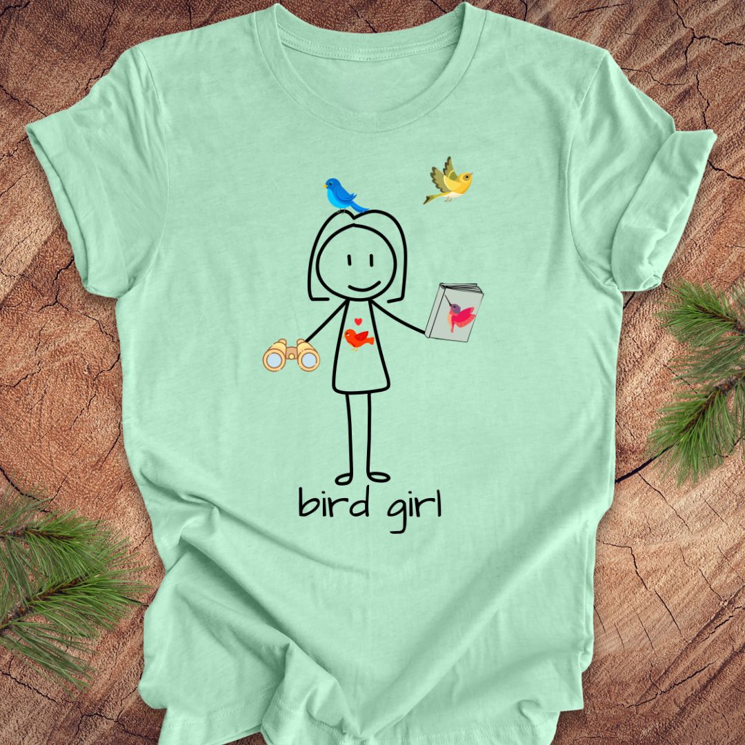 Light green t-shirt with a stick figure design labeled 'bird girl' on a wooden surface.