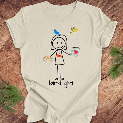 Beige t-shirt with a stick figure design labeled 'bird girl' on a wooden surface.