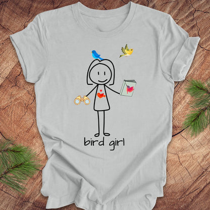 Silver t-shirt with a stick figure design labeled 'bird girl' on a wooden surface.