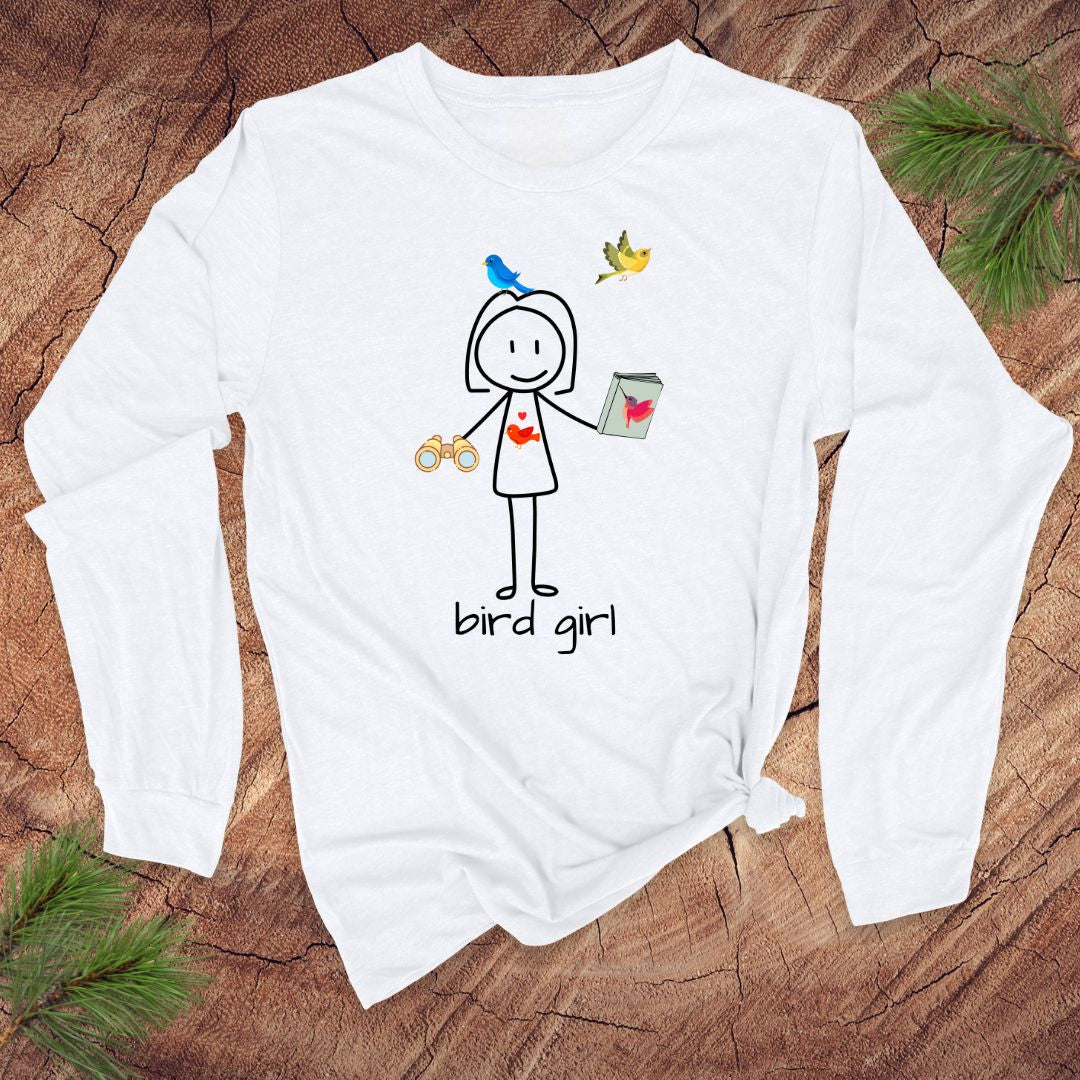 White long-sleeve shirt with 'bird girl' text and stick figure girl with binoculars and guide book, on a wooden surface with pine branches.