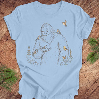 A light blue crewneck t-shirt featuring a graphic of a smiling Yeti surrounded by small yellow and brown birds, displayed flat on a rustic wood background with pine branches.