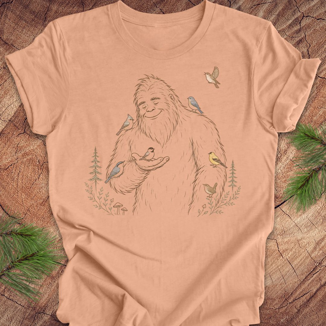 A peach-colored short-sleeve t-shirt with a detailed line art graphic of a Bigfoot surrounded by various colorful birds, laid flat on a rustic wood background with pine branches.