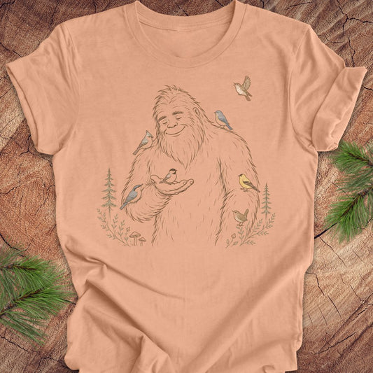 A peach-colored short-sleeve t-shirt with a detailed line art graphic of a Bigfoot surrounded by various colorful birds, laid flat on a rustic wood background with pine branches.