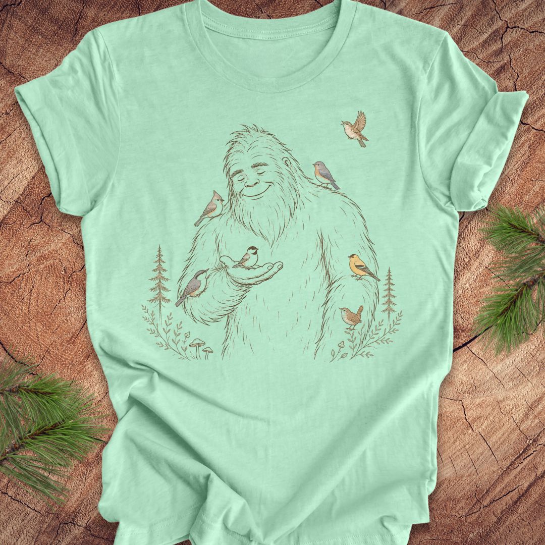 A mint green short-sleeve t-shirt with a detailed graphic of a friendly Bigfoot surrounded by small birds and pine trees, laid flat on a rustic wood background with pine branches in the corners.