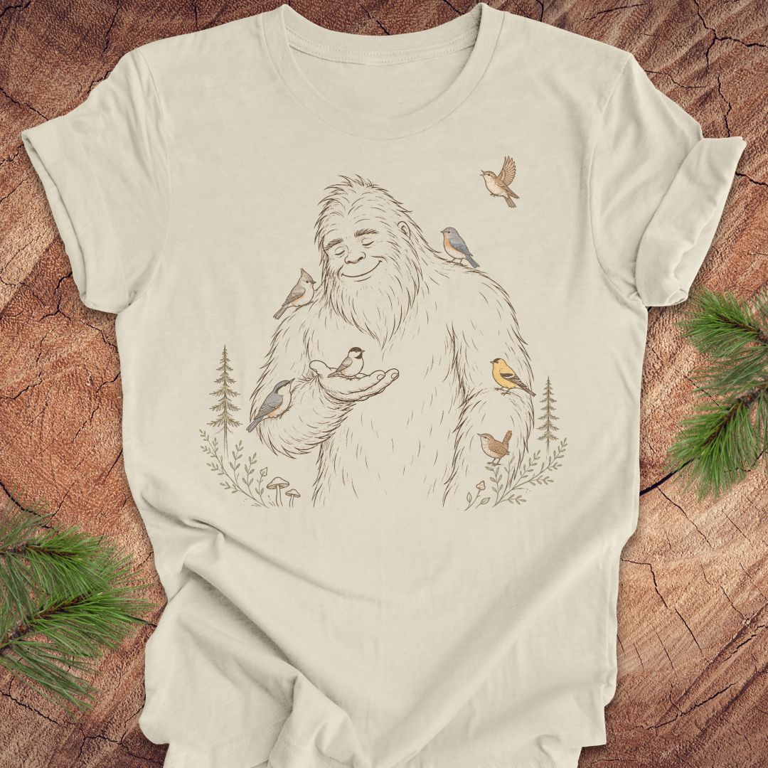 An off-white t-shirt featuring a detailed graphic of a friendly Yeti surrounded by small birds and pine trees, laid flat on a rustic wood background with green pine branches in the corners.