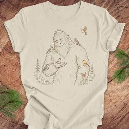 An off-white t-shirt featuring a detailed graphic of a friendly Yeti surrounded by small birds and pine trees, laid flat on a rustic wood background with green pine branches in the corners.