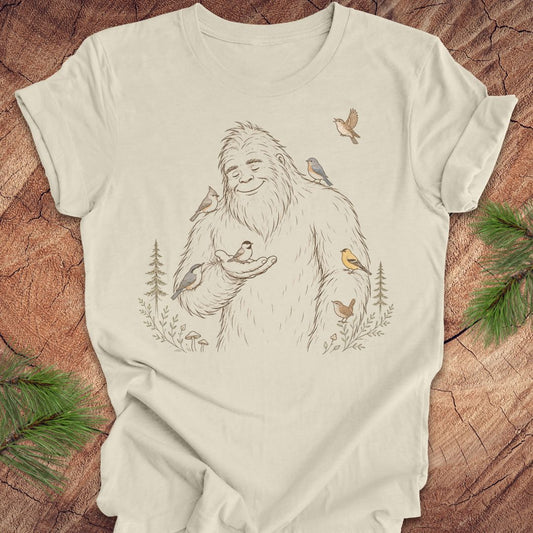 An off-white t-shirt featuring a detailed graphic of a friendly Yeti surrounded by small birds and pine trees, laid flat on a rustic wood background with green pine branches in the corners.