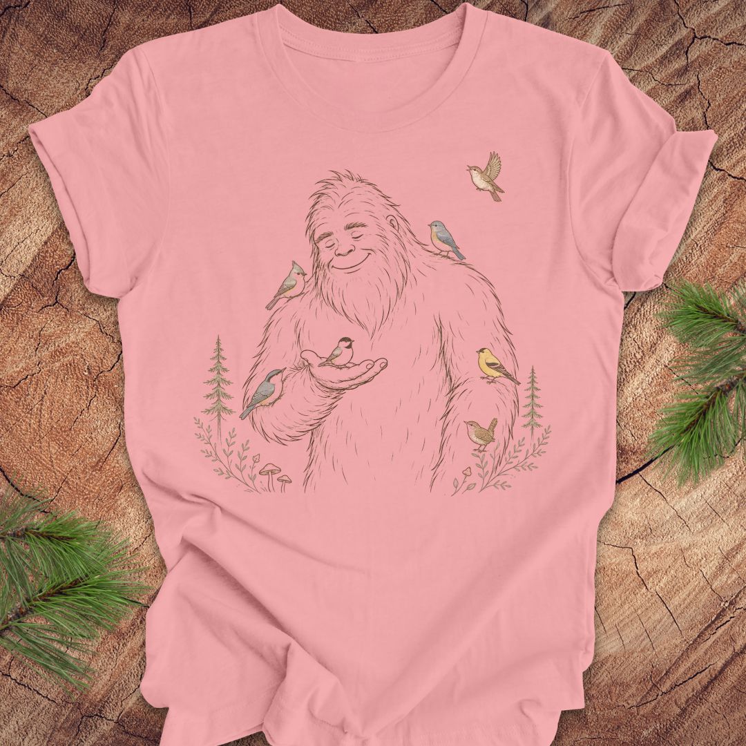 A pink short-sleeve t-shirt with a detailed graphic of a friendly Bigfoot surrounded by small birds and pine trees, laid flat on a rustic wood background with pine branches in the corners.