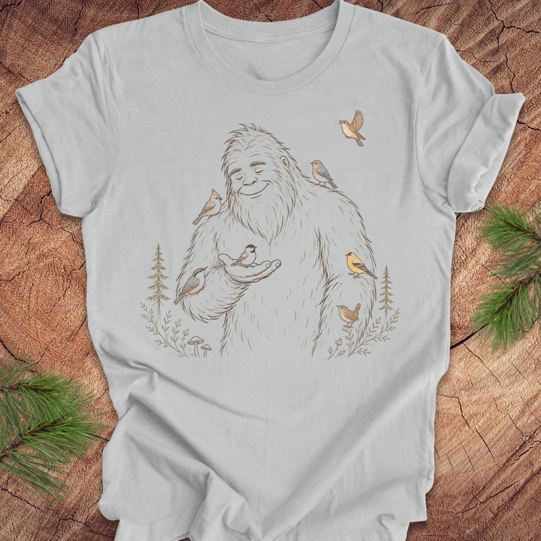 A light gray crewneck t-shirt featuring a line art graphic of a friendly Yeti surrounded by small birds and pine trees, laid flat on a rustic wood background with pine branches.