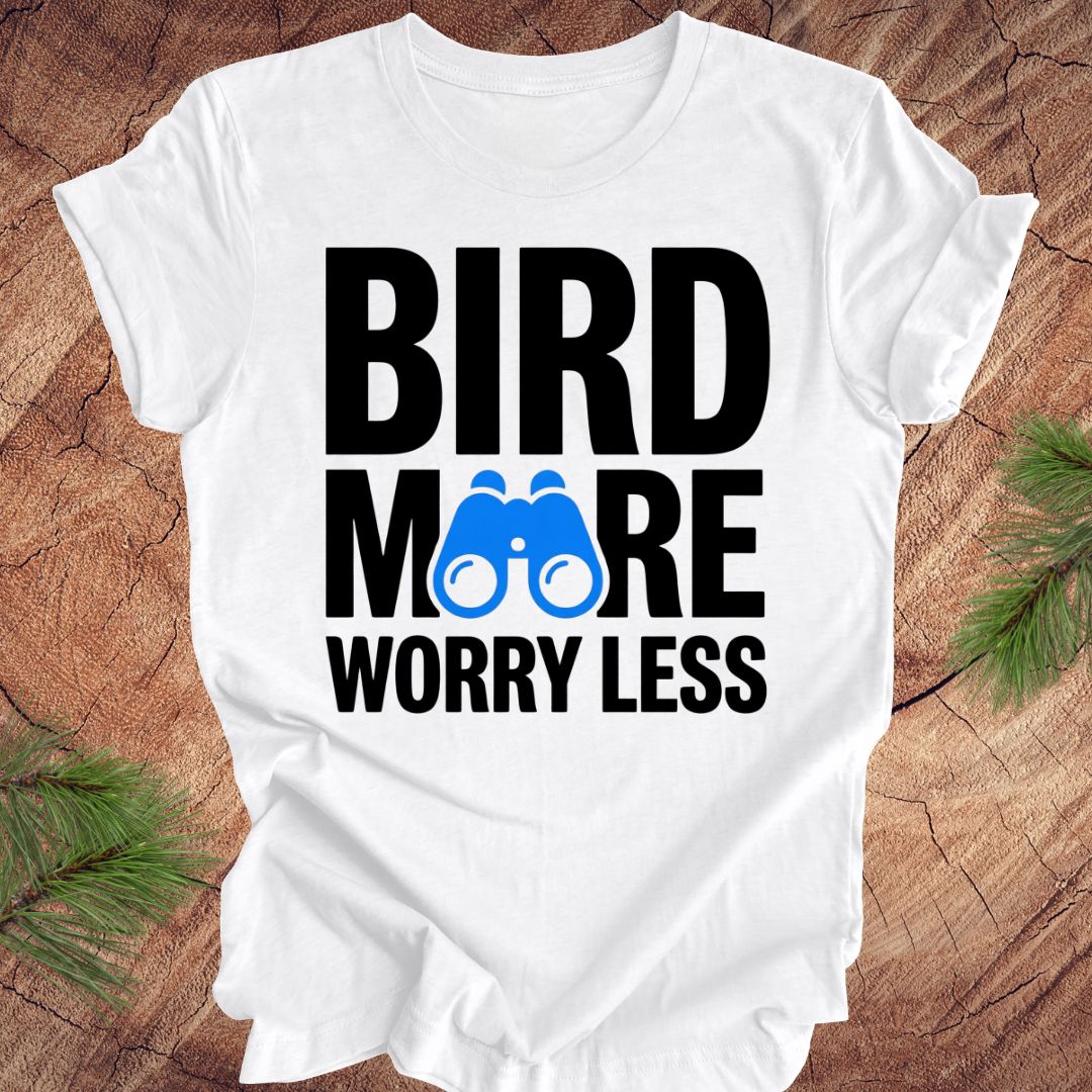 White tshirt with 'BIRD MORE WORRY LESS' text and binoculars on a wooden surface with greenery.