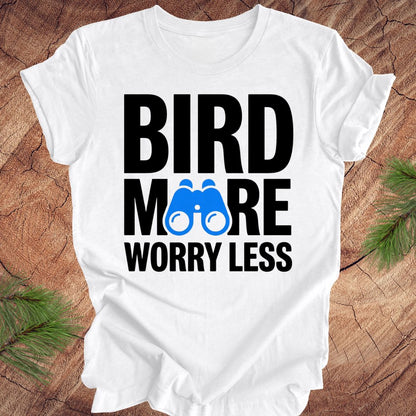 White tshirt with 'BIRD MORE WORRY LESS' text and binoculars on a wooden surface with greenery.