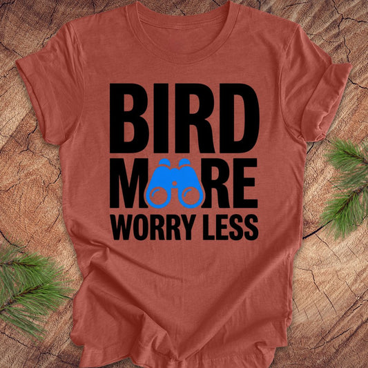 Clay colored tshirt with 'BIRD MORE WORRY LESS' text and binoculars on a wooden surface with greenery.