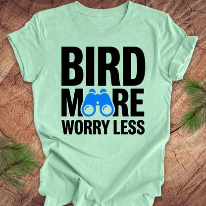 Mint green tshirt with 'BIRD MORE WORRY LESS' text and binoculars on a wooden surface with greenery.