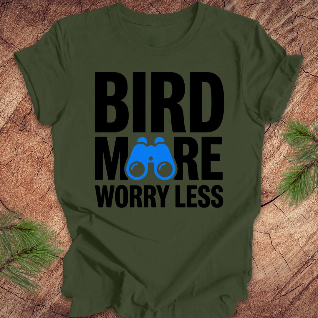 Green tshirt with 'BIRD MORE WORRY LESS' text and binoculars on a wooden surface with greenery.