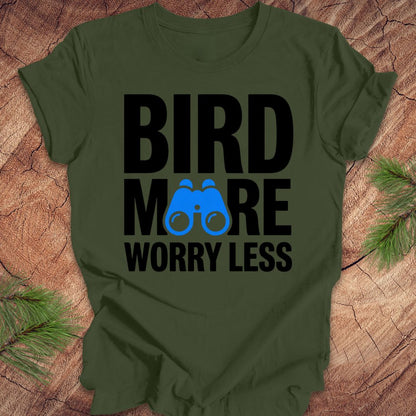 Green tshirt with 'BIRD MORE WORRY LESS' text and binoculars on a wooden surface with greenery.