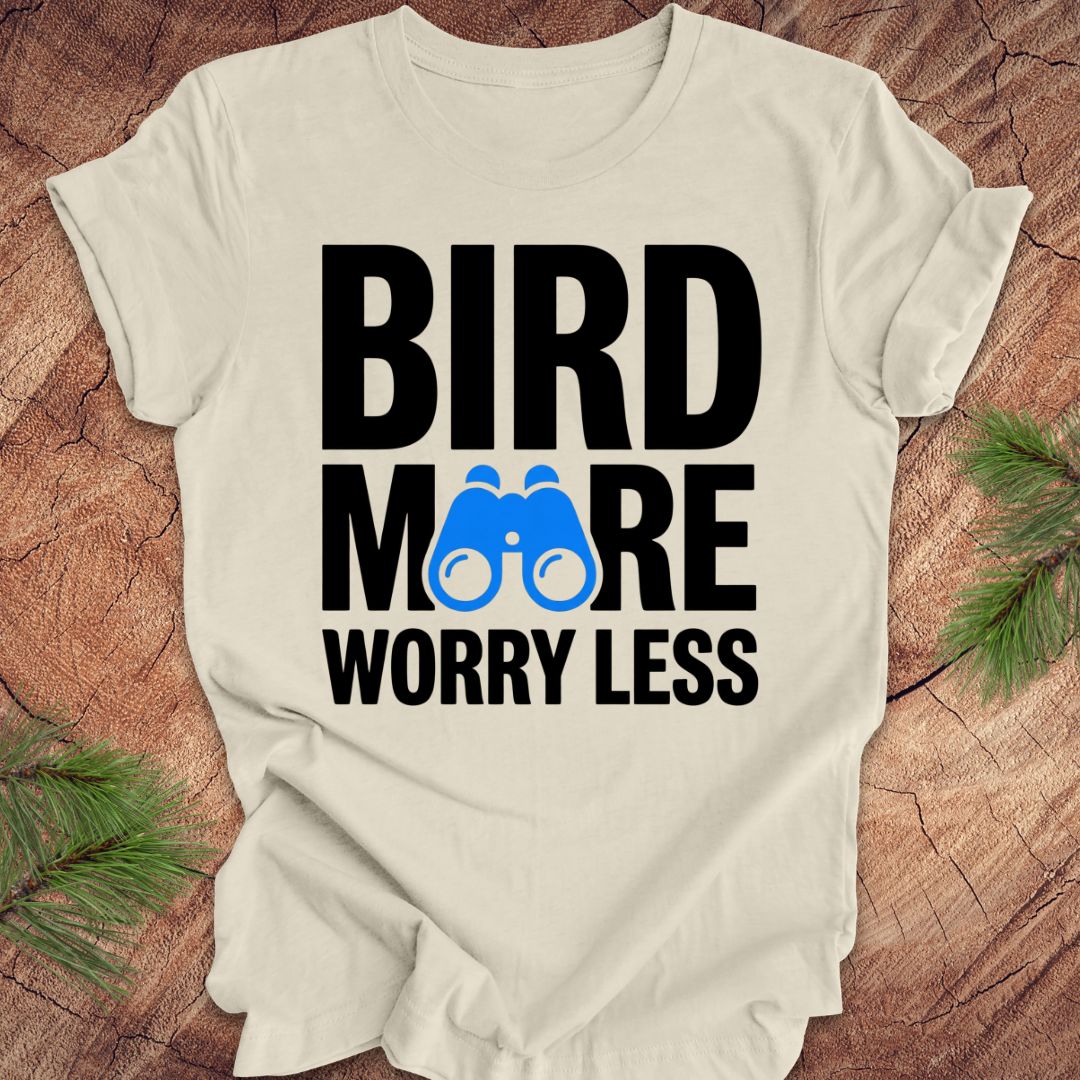 Beige tshirt with 'BIRD MORE WORRY LESS' text and binoculars on a wooden surface with greenery.