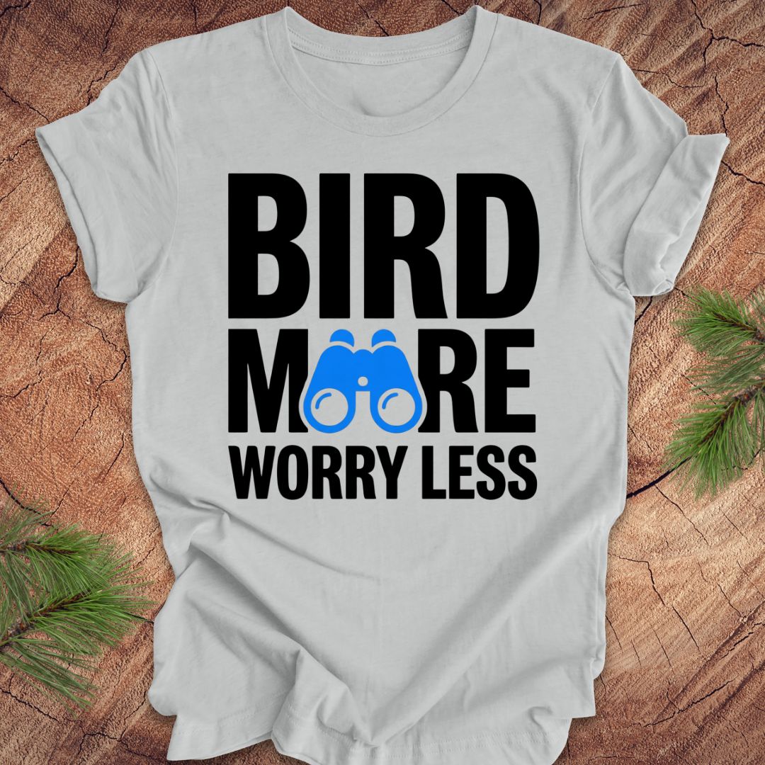Gray tshirt with 'BIRD MORE WORRY LESS' text and binoculars on a wooden surface with greenery.