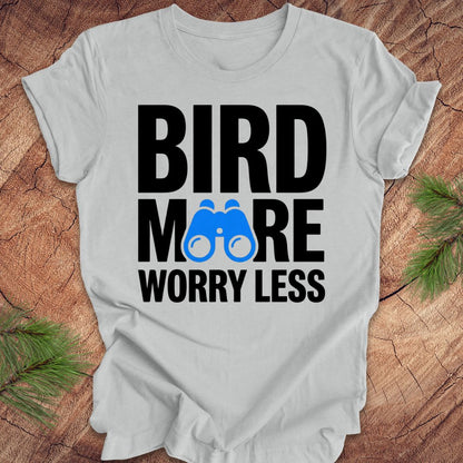 Gray tshirt with 'BIRD MORE WORRY LESS' text and binoculars on a wooden surface with greenery.