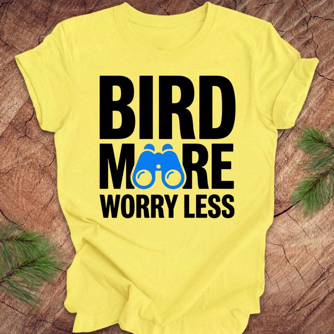 Yellow tshirt with 'BIRD MORE WORRY LESS' text and binoculars on a wooden surface with greenery.