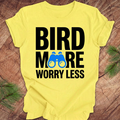 Yellow tshirt with 'BIRD MORE WORRY LESS' text and binoculars on a wooden surface with greenery.