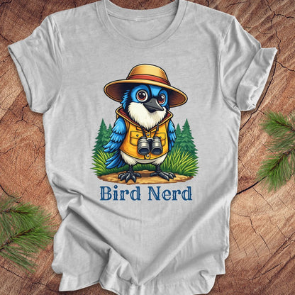 A light gray short-sleeve t-shirt laid flat on a rustic wood background with pine branches in the corners. The shirt features a colorful cartoon graphic of a blue bird wearing a safari hat, yellow vest, and binoculars, with the text 'Bird Nerd' printed below.