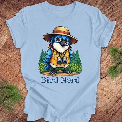 A light blue short-sleeve t-shirt laid flat on a rustic wood background with pine branches at the corners. The shirt features a colorful cartoon graphic of a blue bird wearing a safari hat, yellow vest, and binoculars, with the text 'Bird Nerd' printed below.