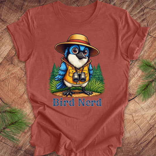 A rust-colored t-shirt featuring a colorful cartoon bird wearing a safari hat and vest with the text 'Bird Nerd' underneath, displayed flat on a rustic wood background with pine branches.