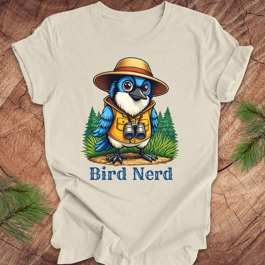 A light beige short-sleeve t-shirt laid flat on a rustic wood background with pine branches in the corners. The shirt features a colorful graphic of a blue bird wearing a safari hat and vest with binoculars, above the text 'Bird Nerd'.