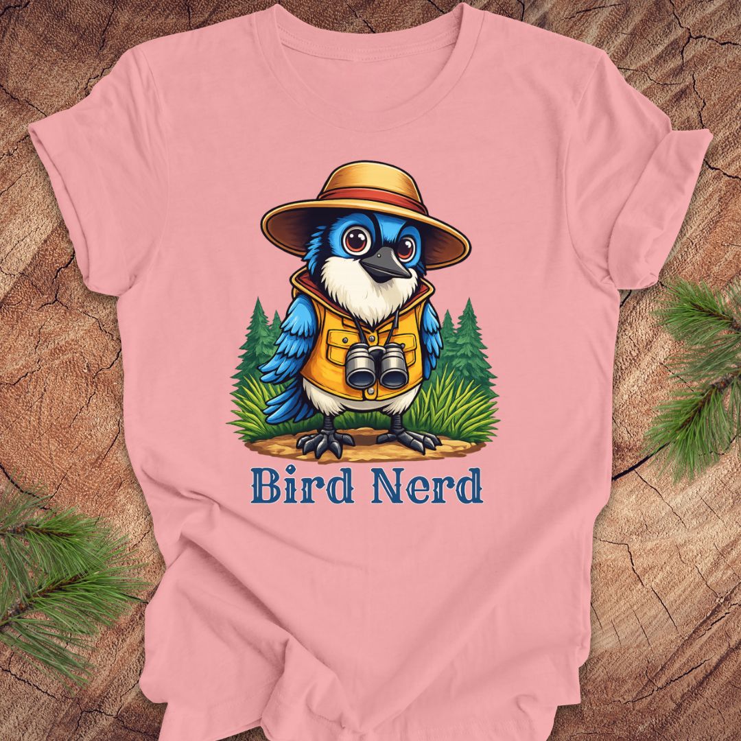 A pink short-sleeve t-shirt laid flat on a rustic wood background with pine branches in the corners. The shirt features a colorful graphic of a blue bird wearing a yellow vest, orange hat, and binoculars, with the text 'Bird Nerd' printed below.