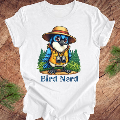 A white short-sleeve t-shirt laid flat on a rustic wood surface, featuring a colorful graphic of a blue bird wearing a safari hat and vest with binoculars, above the text 'Bird Nerd'.