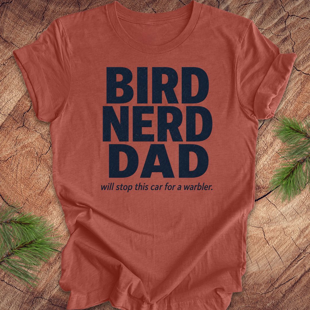 Red t-shirt with 'BIRD NERD DAD' text on a wooden surface with pine branches.