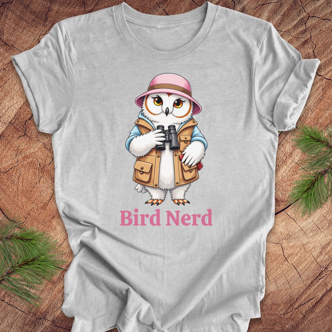 A light gray short-sleeve t-shirt laid flat on a rustic wood background with pine branches in the corners. The shirt features a cartoon owl wearing a pink hat and tan vest while holding binoculars, with the text 'Bird Nerd' printed below in pink.