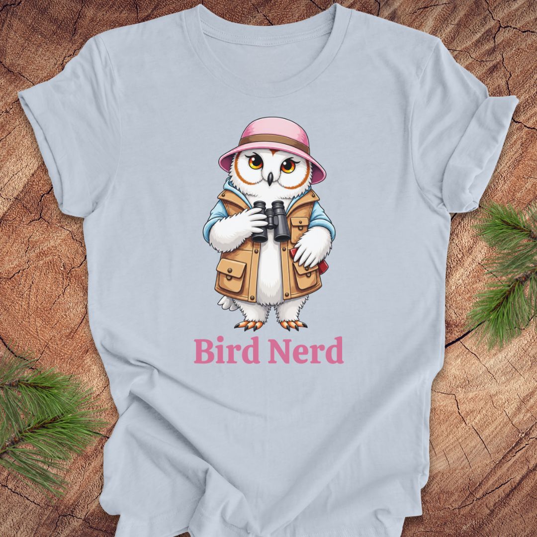 A light blue crewneck t-shirt laid flat on a rustic wood background with pine branches in the corners. The shirt features a cartoon graphic of a white owl wearing a pink hat and tan vest, holding binoculars, with the text 'Bird Nerd' printed below in pink.