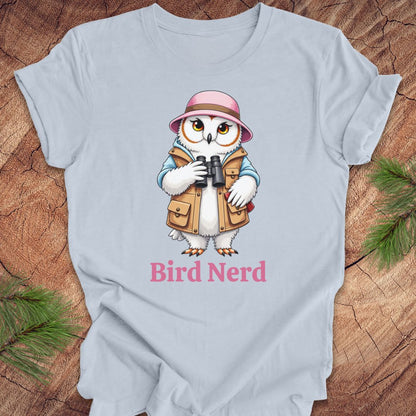 A light blue crewneck t-shirt laid flat on a rustic wood background with pine branches in the corners. The shirt features a cartoon graphic of a white owl wearing a pink hat and tan vest, holding binoculars, with the text 'Bird Nerd' printed below in pink.
