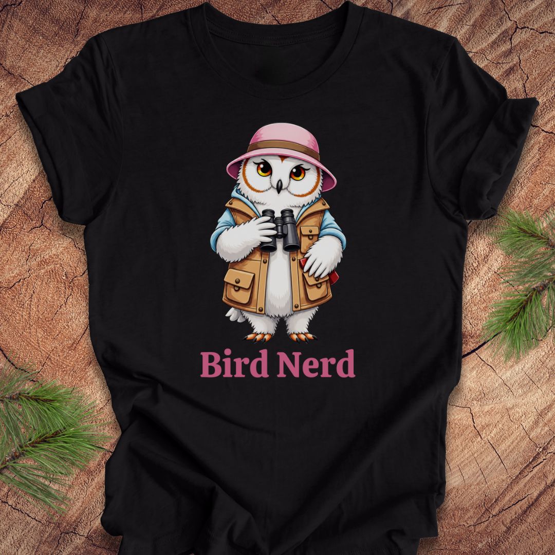A black short-sleeve t-shirt laid flat on a rustic wood background with pine branches in the corners. The shirt features a cartoon owl dressed as a bird explorer wearing a pink hat, blue jacket, and brown vest, holding binoculars, with the text 'Bird Nerd' printed below in pink.