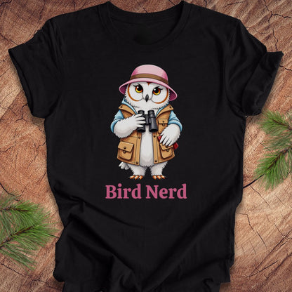A black short-sleeve t-shirt laid flat on a rustic wood background with pine branches in the corners. The shirt features a cartoon owl dressed as a bird explorer wearing a pink hat, blue jacket, and brown vest, holding binoculars, with the text 'Bird Nerd' printed below in pink.
