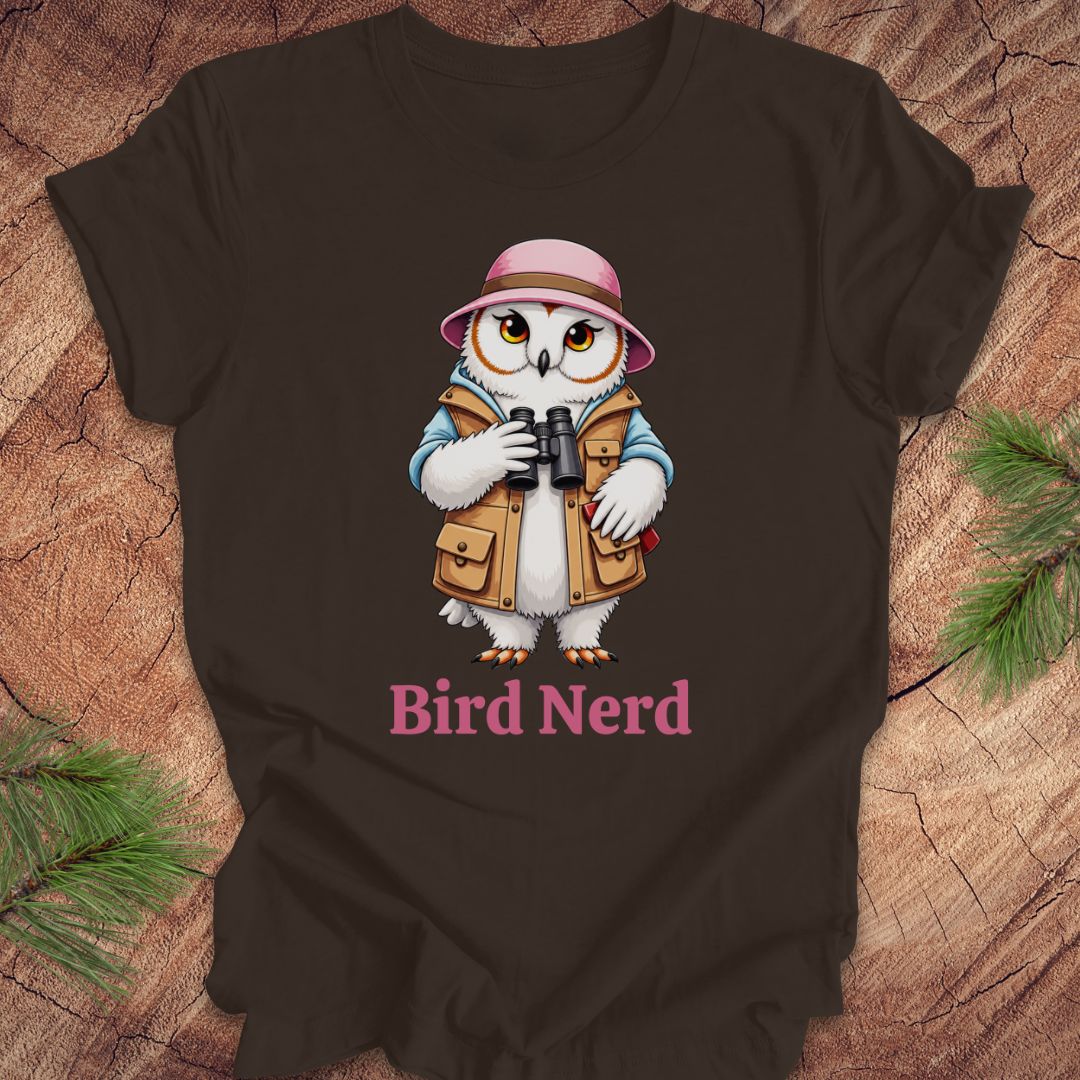 A dark brown t-shirt laid flat on a rustic wood background with pine branches. The shirt features a cartoon graphic of a white owl wearing a pink safari hat and tan vest, holding binoculars, with the text 'Bird Nerd' printed below in pink.