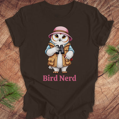 A dark brown t-shirt laid flat on a rustic wood background with pine branches. The shirt features a cartoon graphic of a white owl wearing a pink safari hat and tan vest, holding binoculars, with the text 'Bird Nerd' printed below in pink.