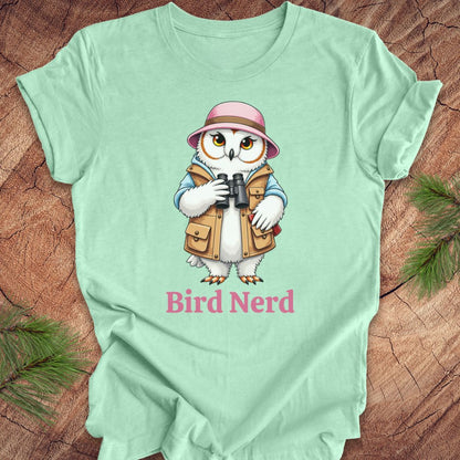 A mint green short-sleeve t-shirt laid flat on a rustic wood background with pine branches in the corners. The shirt features a cartoon owl wearing a pink hat and tan vest, holding binoculars, with the text 'Bird Nerd' printed below in pink.