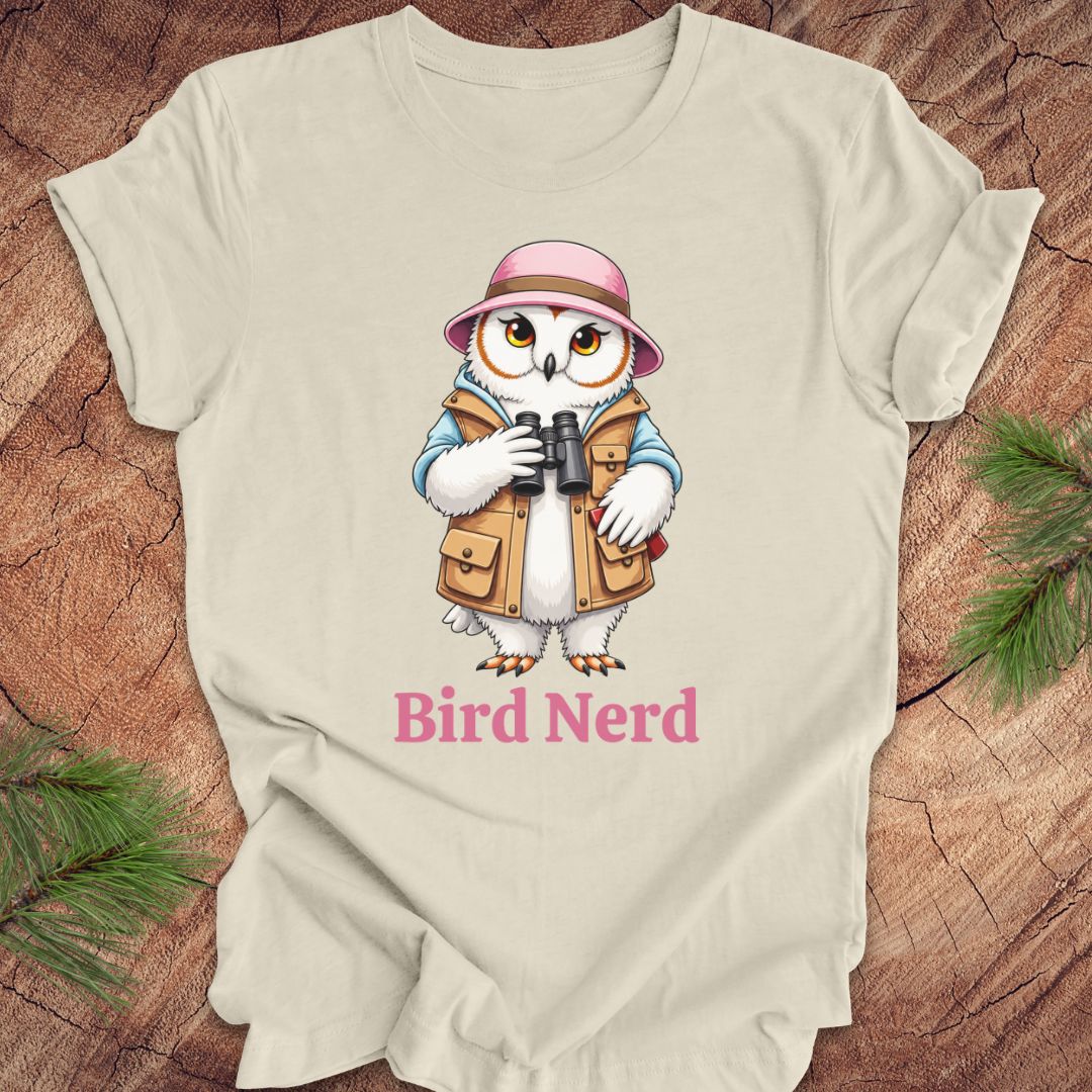 A light beige short-sleeve t-shirt laid flat on a rustic wood background with pine branches in the corners. The shirt features a cartoon graphic of a white owl wearing a pink hat and tan vest, holding binoculars, with the text 'Bird Nerd' printed below in pink.
