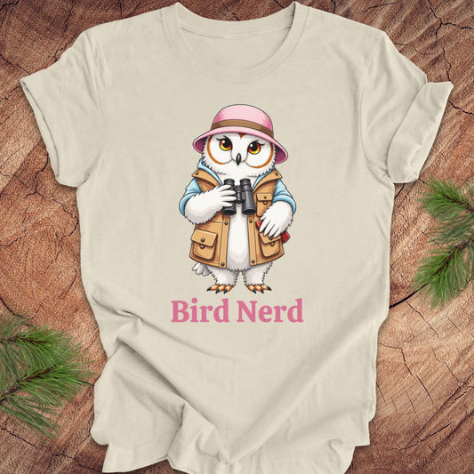 A light beige short-sleeve t-shirt laid flat on a rustic wood background with pine branches in the corners. The shirt features a cartoon graphic of a white owl wearing a pink hat and tan vest, holding binoculars, with the text 'Bird Nerd' printed below in pink.