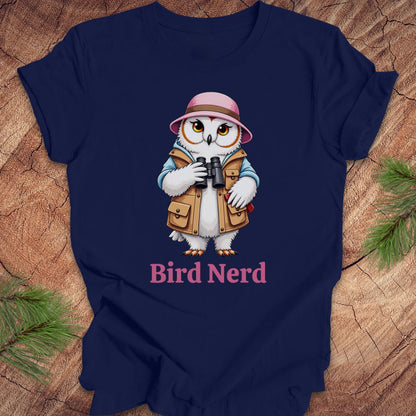 A navy blue short-sleeve t-shirt laid flat on a rustic wood background with pine branches in the corners. The shirt features a cartoon owl wearing a pink hat and tan vest while holding binoculars, with the text 'Bird Nerd' printed below in pink.