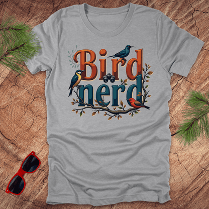 An athletic heather t-shirt with a graphic print of birds and the text 'Bird nerd', surrounded by foliage and a pair of sunglasses lying next to it on a wooden surface.