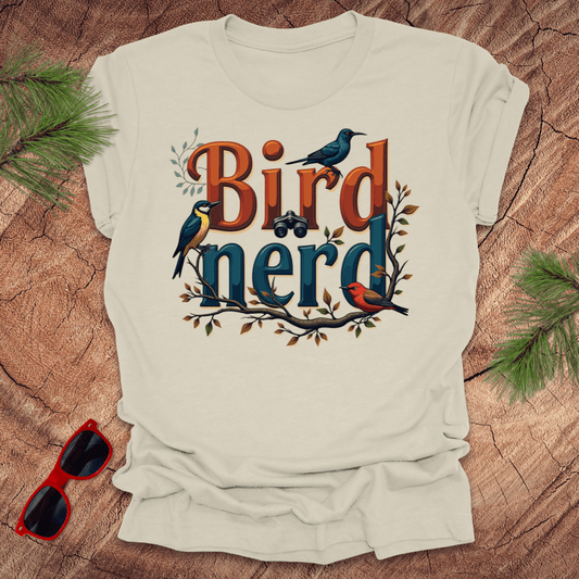 A beige t-shirt with a graphic print of birds and the text 'Bird nerd', surrounded by foliage and a pair of sunglasses lying next to it on a wooden surface.