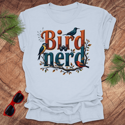 A light blue t-shirt with a graphic print of birds and the text 'Bird nerd', surrounded by foliage and a pair of binoculars lying next to it on a wooden surface.